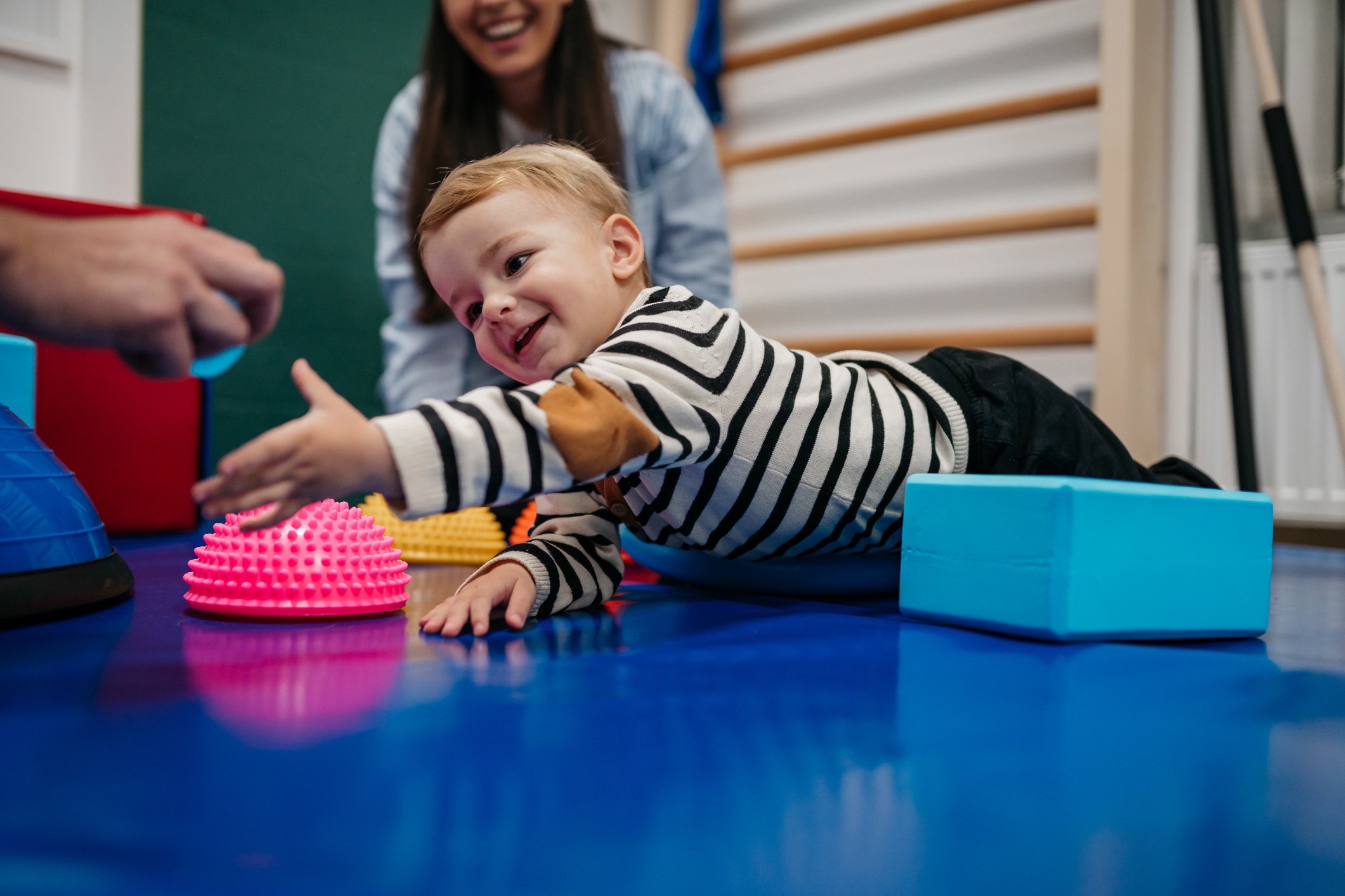 Kleiner Junge liegt auf dem Bauch und greift nach etwas. Hand die ihn etwas hinhält sind noch am Rand zu sehen. eine Frau sitzt im Hintergrund - Bobath Grundkurs.